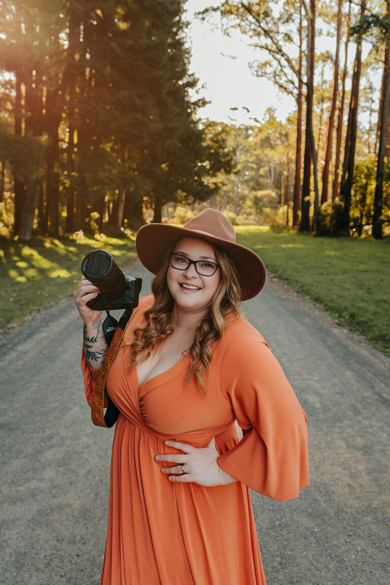 Emma Baker Melbourne Photographer Woman in Orange long sleve dress brown hat blonde hair holding canon camera golden light behind tall trees behind 768x1151